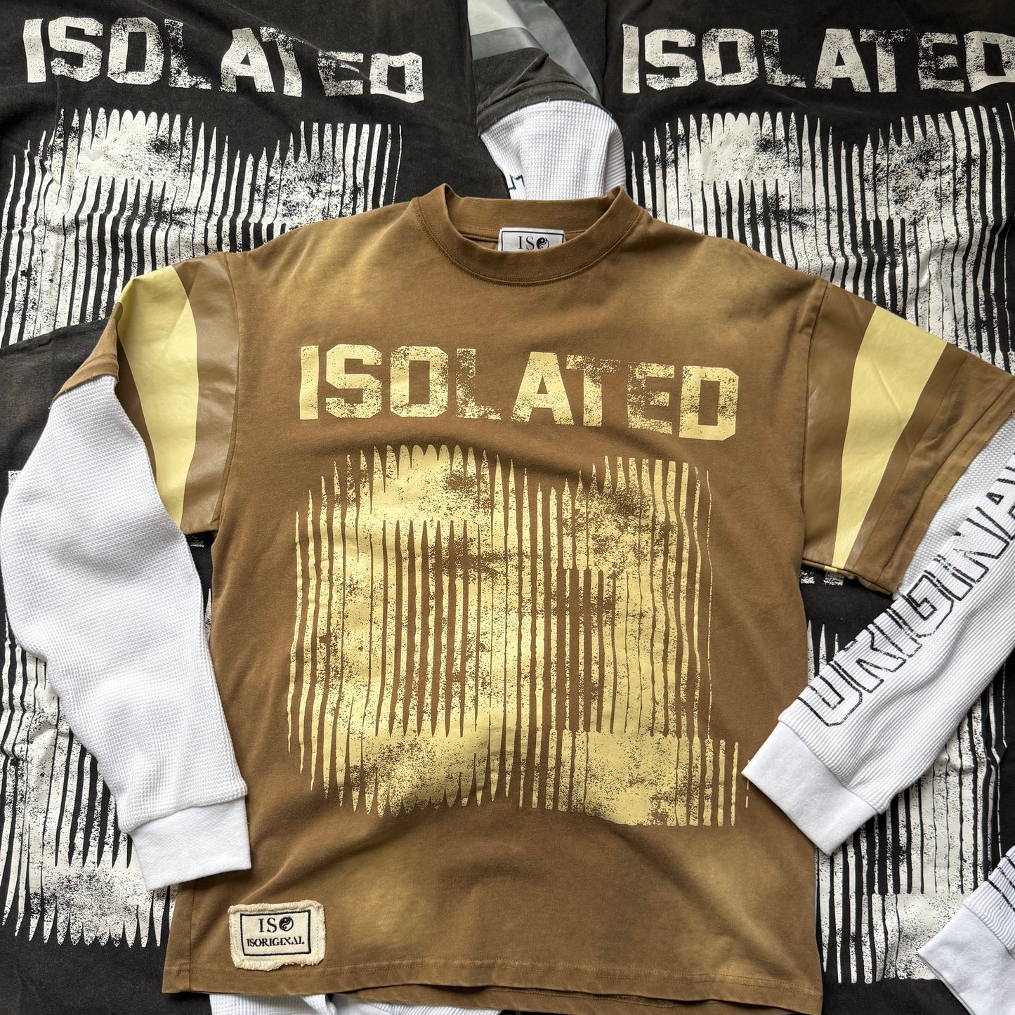 A brown graphic t-shirt with a distressed yellow 'ISOLATED' print and yellow shoulder stripes, laid flat on top of other black and white apparel.